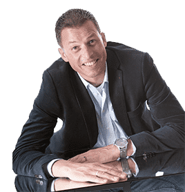 Lachende man in een donker jasje, leunend over een reflecterend oppervlak, met een wit overhemd en horloge.